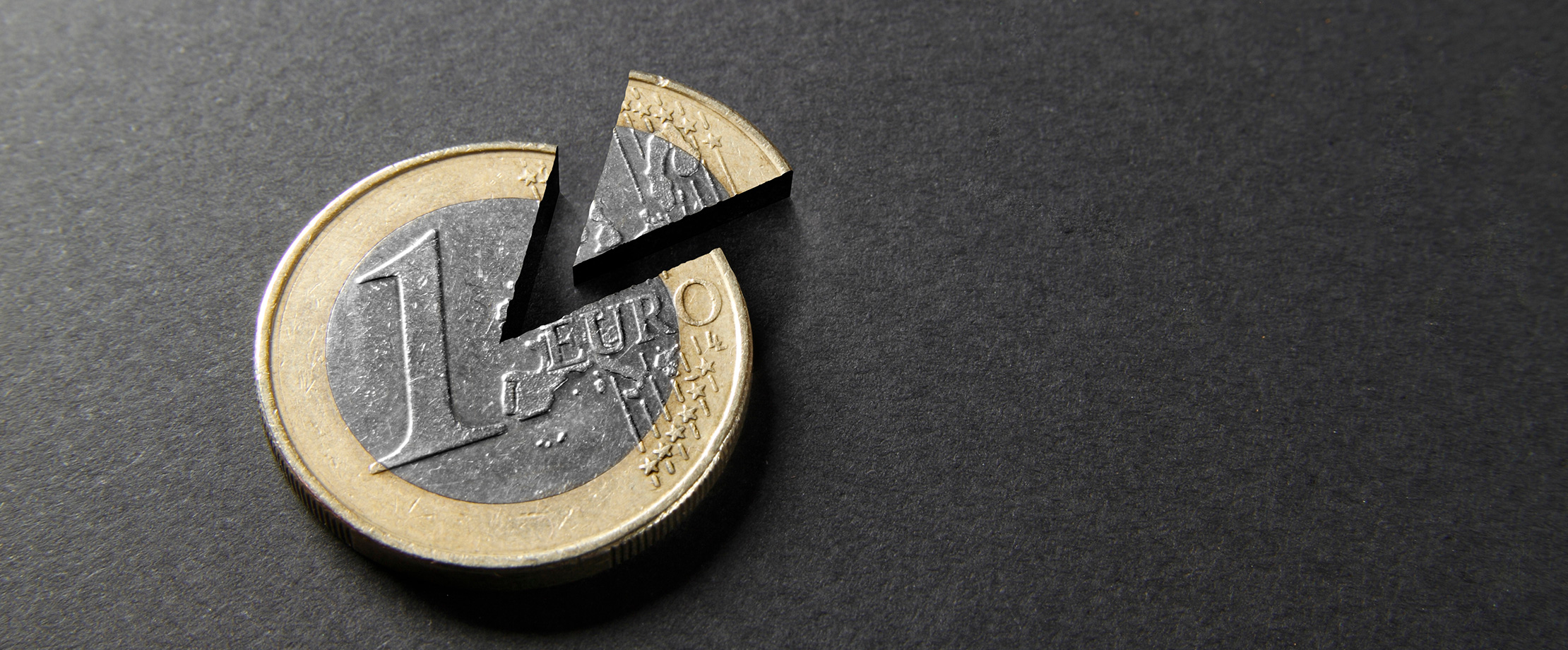 Inflation Ein 1-Euro-Sück liegt auf einer schwarzen Tischplatte. Ein kuchenförmiges Stück wurde aus dem Euro ausgeschnitten und liegt abgetrennt daneben.
