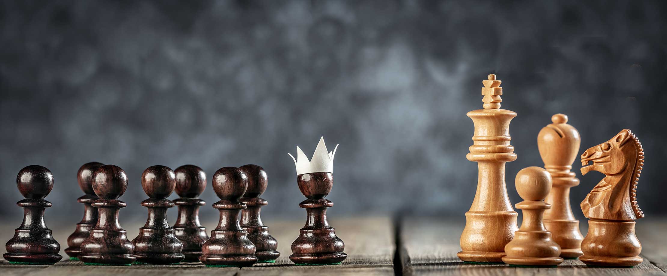 Ihr Vermögensberater & Kreditmakler Schachfiguren stehen auf einem Tisch vor schwarzem Hintergrund. Die weißen Figuren rechts sind der König, ein Läufer, Ein Pferd und ein Bauer. Die schwarzen Figuren links sind alle Bauern, der vorderste von ihnen trägt eine Papierkrone.