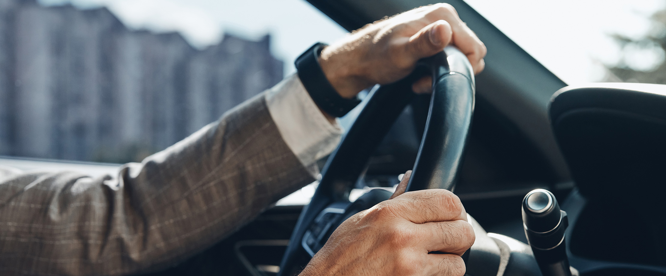 Sanierung des Kfz-Versicherungsvertrages Ein Mann im Anzug fährt mit dem Auto. Man kann seine Arme und Hände sehen, mit denen er das Lenkrad hält. Am Handgelenk trägt er eine schwarze Armbanduhr.