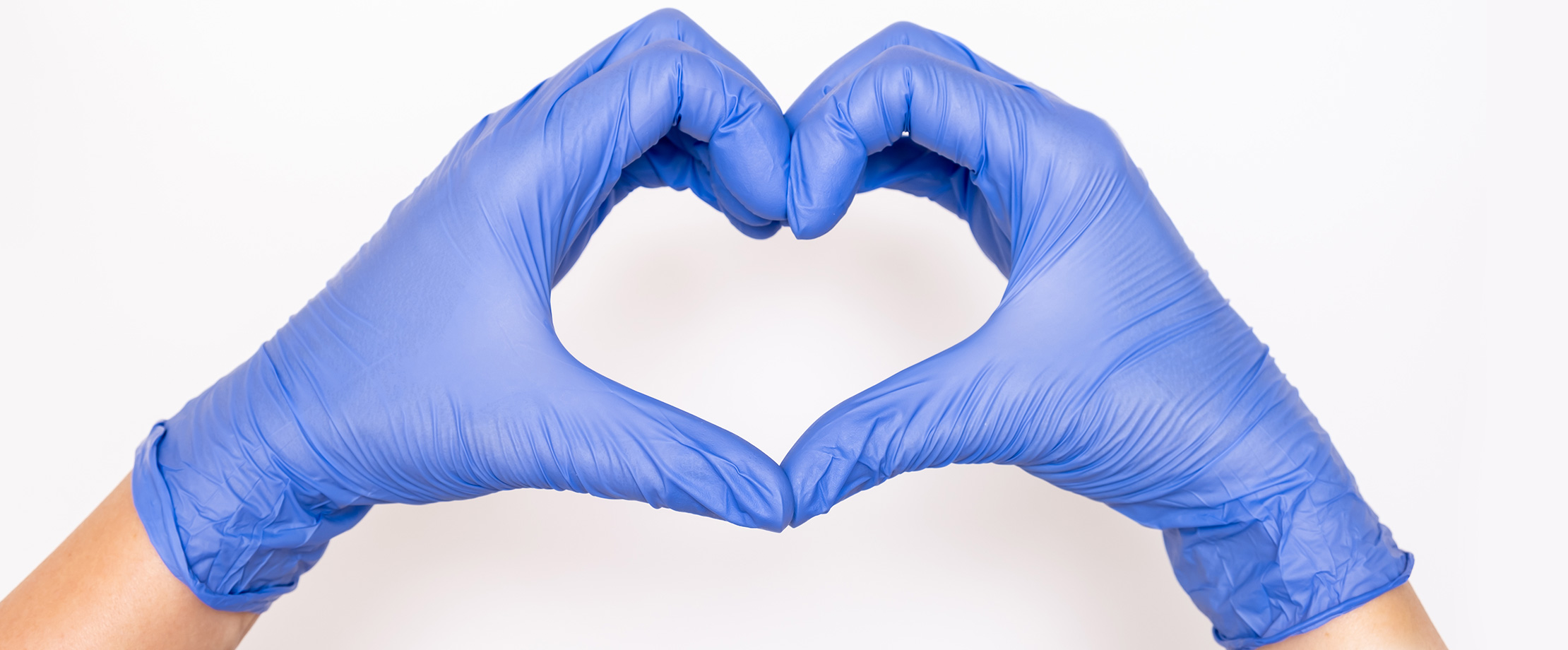 Stationäre Behandlungen, Sonderklasse Zwei Hände in blauen Chirurgenhandschuhen formen das Love-Sign. Dieses blaue, behandschuhte Herz ist vor einem sterilen weißen Hintergrund zu sehen.