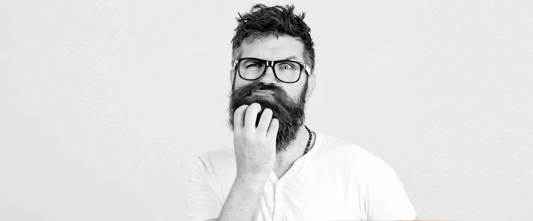 Gesetzliche & private Krankenversicherung Ein Mann mit Brille und Bart greift sich ans Kinn und verzieht nachdenklich das Gesicht. Es ist ein Schwarz-weiß-Bild. Der Blick des Mannes ist prüfend und auch etwas besorgt.