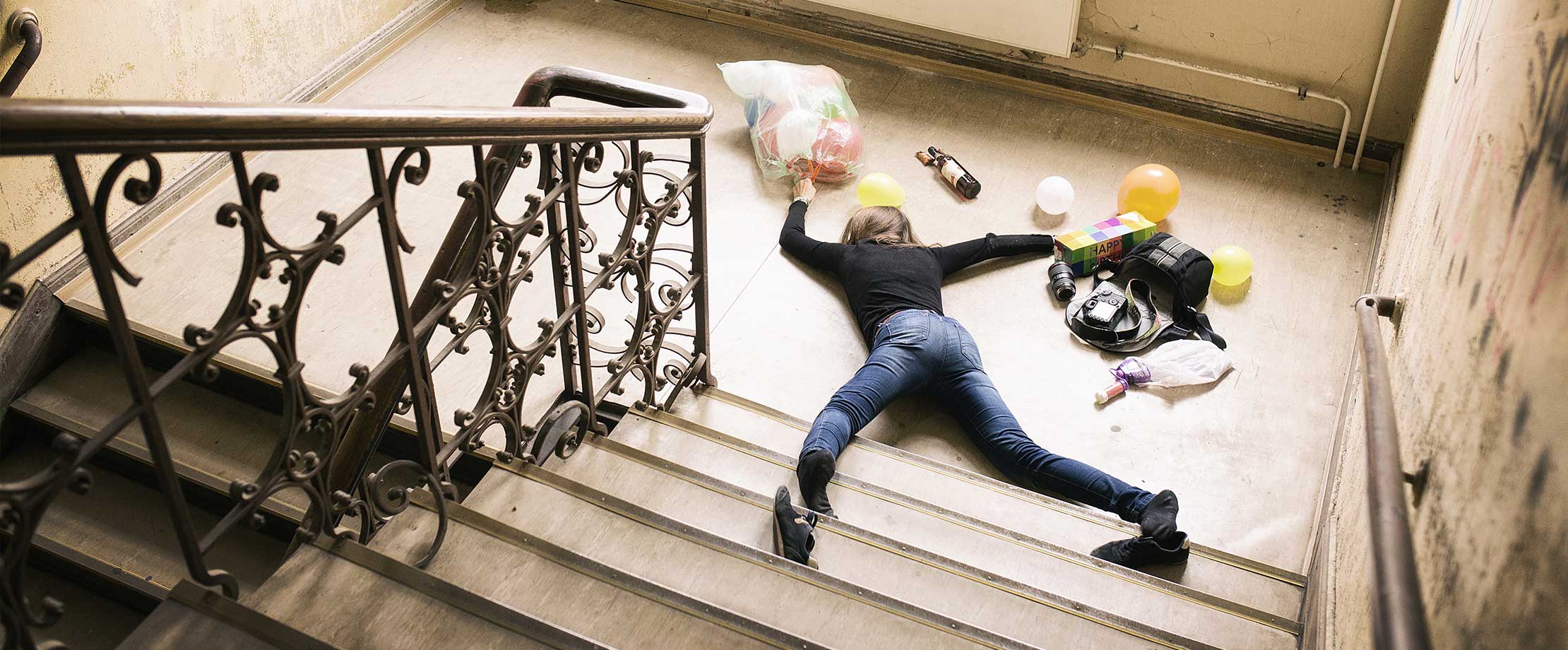 Gesetzliche & private Unfallversicherung Eine Frau liegt am Fuß einer Treppe, nachdem sie gestürzt ist. Um Sie herum liegen Einkäufe, ihre Tasche, eine Kamera und ein Geburtstagsgeschenk.